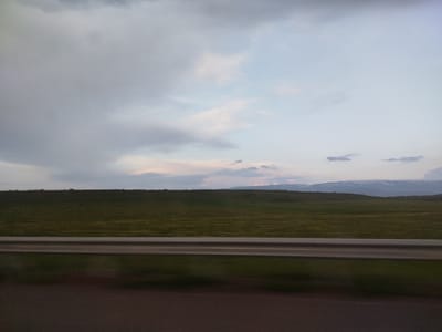The image captures a rural landscape, possibly in the vicinity of Sarnaghbyur, Armenia, under an overcast sky with patches of pale blue and soft pink clouds. A vast, undulating field of green grass stretches across the midground, its texture suggesting a slight breeze. In the distance, a faint outline of hills or mountains can be discerned, with some areas appearing snow-capped. A metallic guardrail, likely separating a road from the field, runs horizontally across the lower third of the image, its polished surface reflecting the muted light. The foreground is dominated by a dark, blurred surface, suggesting a road or asphalt. There are no people or distinct objects within the frame, focusing the viewer's attention on the natural environment.