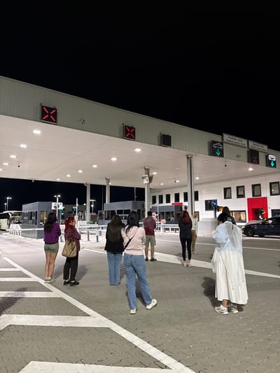 A group of people are standing at the border crossing of Gostun, in the Prijepolje Municipality, Serbia, on the border between Serbia and Montenegro. The building has white walls with lighted “X” signs and green arrows for the customs lanes. The people are waiting in a paved area with white lines. It is nighttime.