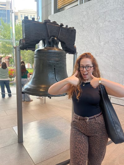 A young adult female is depicted standing indoors in the Liberty Bell Center, Philadelphia, United States. She is positioned to the right of the historic Liberty Bell, a large bronze artifact with a prominent vertical crack and visible inscriptions, mounted on a dark support frame. The female has wavy reddish-brown hair and wears dark-rimmed glasses, a black sleeveless top, brown leopard-print wide-leg pants with a brown belt, a gold-toned wristwatch, and carries a black shoulder bag on her left arm. She smiles broadly with an open mouth, extending both arms forward with thumbs raised in a "thumbs up" gesture. Behind the Liberty Bell, through large glass windows, green trees, historical brick structures, and modern glass skyscrapers are visible in the outdoor cityscape. Three other individuals are partially visible in the left background, with their backs to the camera, observing the exterior view. The interior space features light-colored marble-patterned wall panels and light-gray tiled flooring.