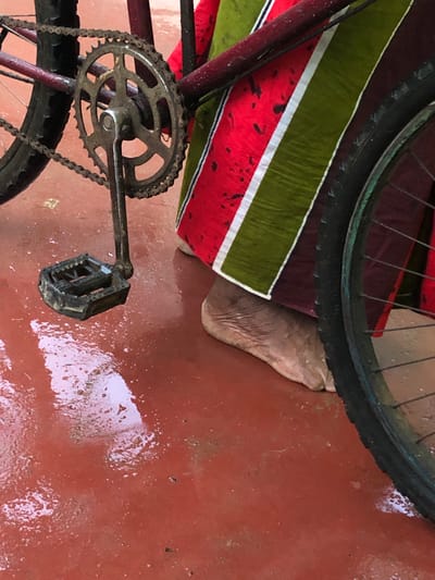 The image shows a close-up of a bicycle's crankset, pedal, and rear wheel, with a portion of a person's foot visible. A colorful cloth, with stripes of red, green, and white, is draped over part of the bicycle frame and the person's foot. The bicycle is resting on a wet, reddish-brown surface, possibly a concrete floor, with several puddles and reflections indicating recent rain. The lighting suggests it might be daytime, with natural light casting shadows and reflections. The scene appears to be outdoors, possibly in a residential area or workshop in Kodagoda, Sri Lanka, given the typical appearance of such surfaces and the presence of a bicycle. There is no visible text in the image.