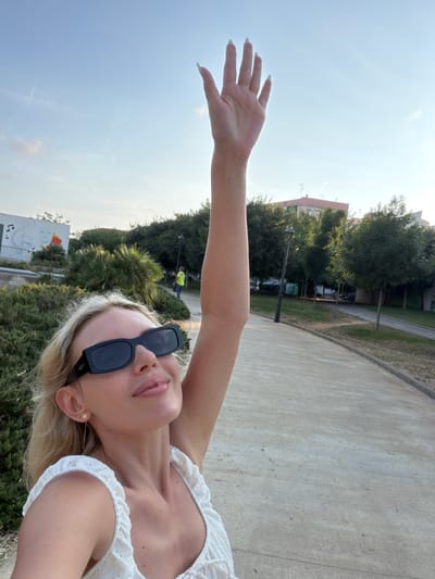 In this selfie, a blonde woman with fair skin and light makeup is shown outdoors. She is wearing a white, eyelet-trimmed top and dark-rimmed sunglasses. A delicate star-shaped earring is visible in her left ear. Her right arm is raised, with her hand extended towards the sky. The setting appears to be a park or public outdoor space with a paved pathway, grass, trees, and some buildings in the background. The sky is clear and blue, suggesting a sunny day. In the distance, two figures can be seen, one wearing a yellow vest, possibly indicating construction or maintenance work. The overall mood seems relaxed and joyful, possibly indicative of a pleasant day spent outdoors. The image appears to have been taken during daylight hours, likely in the afternoon given the lighting. The location context of Benimàmet-beniferri, Spain, is provided.