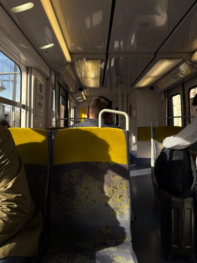 The image shows the interior of a train carriage in Bourg-la-Reine, France. The train is in motion, as indicated by the blurred view outside the windows. The passenger nearest to the camera is a man, wearing a brown jacket, seated in the leftmost seat, facing forward. Behind him is another man, wearing a dark jacket and a baseball cap, seated in the next seat facing forward, and on the right is a person wearing a striped shirt and seated facing the left, towards the window. The train carriage is equipped with overhead lights. There is a large black backpack and a black suitcase on the floor of the train carriage, next to the person wearing a striped shirt. The seats in the train carriage are yellow and gray and upholstered with a patterned fabric.