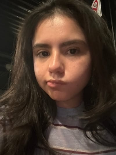A close-up, head-and-shoulders portrait of a female subject with long, dark hair, appearing to be a young adult. Her eyes are dark and look directly at the viewer; her lips are slightly pursed, conveying a neutral expression. She wears a light blue long-sleeved shirt with thin, horizontal maroon stripes. The background is dark and out of focus, indicating an indoor setting with a visible industrial-style ceiling structure. A white sign with a red circular outline and a diagonal red line, consistent with a prohibition symbol, is partially visible in the upper right quadrant. The location is in the city of Santiago, Mexico.