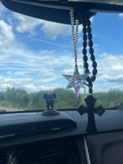The image shows the interior of a car looking out at a cloudy sky and a tree-lined horizon, possibly in the city of Gubachevo, Russia. Hanging from the rearview mirror are three decorative items: a strand of clear beads, a faceted crystal star pendant, and a dark wooden cross. In the foreground, on the dashboard, is a small black figurine of a bear wearing a crown. The car's infotainment screen is visible at the bottom of the frame, displaying text that appears to be song information.
