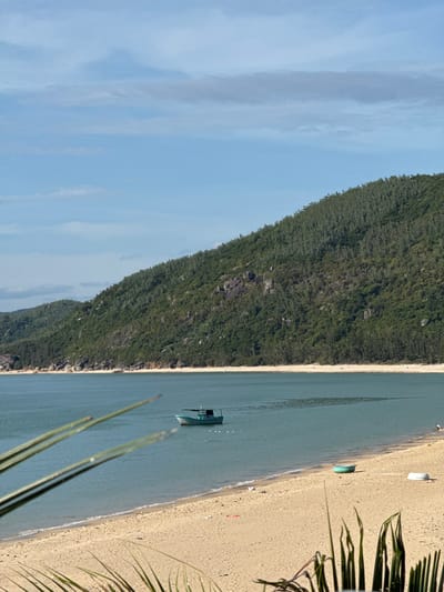 The image depicts a tranquil beach scene in Song Cau, Vietnam. A calm, turquoise body of water stretches towards a lush, green, tree-covered mountainside that forms the backdrop. In the foreground, a wide, sandy beach is visible with a few small boats and what appear to be beach umbrellas or shelters scattered across it. A single, green and blue fishing boat is anchored or drifting in the shallow water near the shore. The sky above is a clear blue with a few wispy clouds. The overall impression is one of a serene, natural coastal environment.
