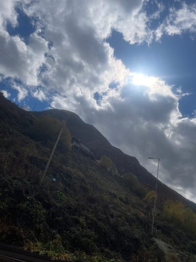 The image captures a steep, vegetated mountain slope in Juta, Georgia, under a partly cloudy sky with bright sunlight. The slope, covered predominantly in dark green and brown foliage, ascends from the lower-left to the upper-right. A multi-story building, likely a guesthouse or lodge with a reddish-brown lower facade and a lighter upper section, is visible horizontally embedded into the mid-upper portion of the slope. Two utility poles stand on the incline: a wooden pole with wires on the left-midground and a metal pole fitted with a street light on the right-midground, also supporting wires that traverse the scene. The upper-right quadrant of the sky features the sun, partially obscured by white and grey clouds, emitting a strong glare and lens flare. Patches of blue sky are visible between the clouds. No individuals or animals are present.