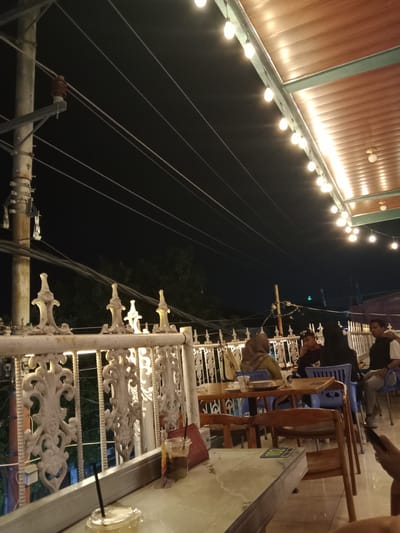 The image depicts an outdoor dining area at night in Pandean, Indonesia. Several people are seated at tables, engaged in dining and conversation. The setting features a white ornate metal railing, wooden tables and chairs, and string lights illuminating the area from above. Electrical wires and a utility pole are visible in the background, along with trees and the night sky. The foreground shows a table with drinks.