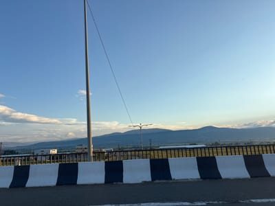 The image depicts a view from a bridge or overpass in the city of Dzveli Kanda, Georgia. In the foreground, a concrete barrier with black and white diagonal stripes serves as a safety railing. Beyond the barrier, a black metal fence encloses a grassy area. In the middle ground, several buildings with white and yellow facades are visible, along with a street lamp. A tall, thin pole, possibly a utility pole, extends vertically on the left side of the frame, with a thin wire running diagonally across the blue sky. The background features a range of hazy, blue-hued mountains under a bright, partly cloudy sky. Two small dark shapes, possibly birds, are visible in the upper right portion of the sky. The overall scene suggests a daytime view of a semi-urban or industrial area on the outskirts of a city, with natural landscapes in the distance.