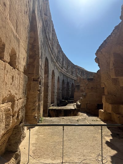 The image depicts a low-angle view inside the Roman amphitheater of El Jem, Tunisia. The scene showcases a fragment of the amphitheater's multi-tiered structure, constructed from light-colored stone. The sky is visible at the top left, contrasting with the dark recesses of the building's arches and openings. A metal support pole and electrical wiring are present on the right side. The architecture displays the characteristic rounded arches and layered stonework of Roman amphitheaters.