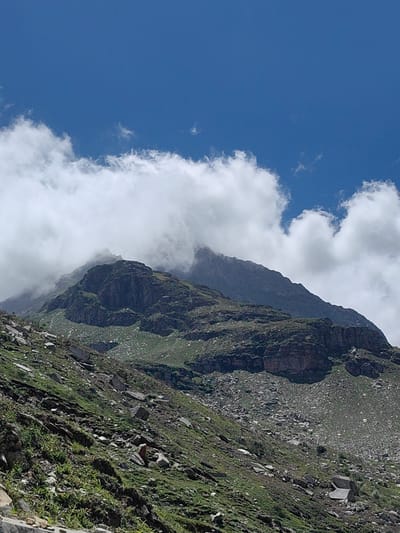A mountainous landscape in KOKSAR, India, features rugged, rocky slopes covered with sparse green vegetation. The foreground shows a steep incline with scattered rocks and patches of grass. In the midground, a prominent rocky ridge with layered strata is visible, partially obscured by lush green grass. The background is dominated by a large mountain, its peak shrouded in swirling white clouds that contrast against a bright blue sky. The overall scene depicts a natural, undeveloped environment under a clear but cloudy sky.