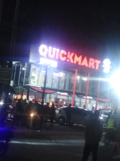 The image depicts a vibrant night scene in Oloosirkon/Sholinke ward, Kenya, centered around a brightly illuminated multi-story building. A large, prominent red neon sign across the top of the building clearly reads "QUICKMART." Below this, on a lower section of the building, another smaller sign with the text "GENTRY" is partially visible.
The building itself features a modern design with numerous large windows and distinct red vertical accents, possibly support columns or decorative panels. The interior of the lower levels appears lit, suggesting activity inside.
In front of the Quickmart building, a significant crowd of people is gathered on the street and sidewalk. Their figures are mostly silhouetted against the bright lights of the building and the surrounding area. Several vehicles, including cars and what appears to be a motorcycle (identified by a bright headlight beam), are present on the road, indicating a busy urban environment.
The time of day is night, as evidenced by the dark sky and the strong reliance on artificial lighting. Bright light sources, possibly streetlights or vehicle headlights, create noticeable lens flares, particularly on the right side and in the lower-center of the image, contributing to a slightly hazy or glowing effect. A small, decorated object, possibly a Christmas tree, can be faintly discerned near the bottom right of the building, hinting at the time of year. The overall impression is one of a bustling commercial or community hub active after dark. The image has a somewhat blurry quality, particularly for distant details and individual faces.