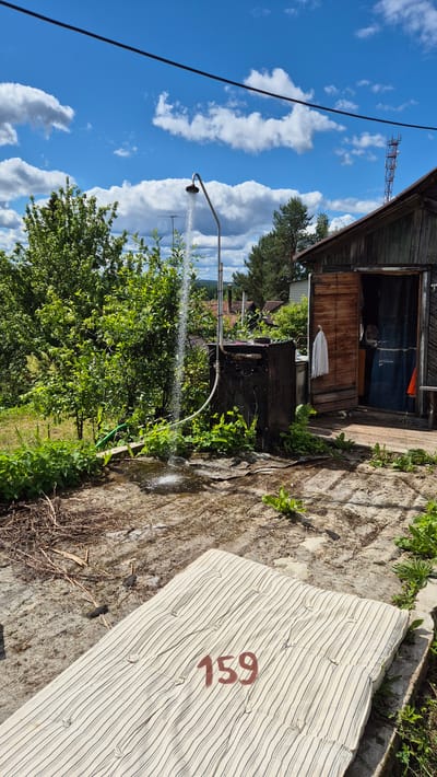 The image depicts an outdoor shower setup next to a rustic wooden structure. Water is actively spraying from a showerhead attached to a curved metal pipe. The showerhead is positioned over a dark, rectangular tank, which appears to be part of the shower system, possibly a water heater or storage. A hose snakes from the tank to the showerhead. Green vegetation, including bushes and trees, surrounds the shower area and the wooden building. The building has an open doorway revealing a blue curtain inside and a white towel hanging on the exterior. In the foreground, a striped mattress with the number "159" stenciled in red is placed on a concrete or stone patio. The ground is a mix of concrete, dirt, and dry debris. The sky above is blue with scattered white clouds, and a power line stretches across the top of the frame. A cell tower is visible in the distance. The scene is set in a rural environment, consistent with a location in Машезеро, Russia, suggesting a simple, utilitarian setup for outdoor living or bathing.