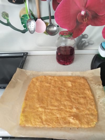 The image shows a flat, golden-brown baked item, possibly a cake or cookie, resting on a sheet of parchment paper. The parchment paper is on top of a dark baking tray. In the background, there's a glass jar containing a dark red liquid with small red berries. Above the jar, kitchen utensils, including spoons and a ladle, are hanging. The wall behind the objects features a large, vibrant floral print, likely an orchid. The setting appears to be a kitchen counter. The location is specified as Andzhero-Sudzhensk, Russia.