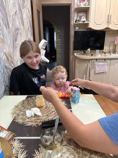 The image depicts a scene in a kitchen, likely in Berëzovka, Russia. Two people are present: a younger girl in the foreground, wearing a black t-shirt with cartoon characters, and a baby in the background, wearing a pink shirt. The baby appears to have food around its mouth. A person's arms, dressed in a blue t-shirt, are visible reaching towards the baby, holding a juice box and a toy. The setting appears to be a dining area within the kitchen, with a table covered by a patterned placemat and a glass of liquid. There are kitchen cabinets and a television visible in the background. The overall mood seems casual and domestic. The time of day and weather are not discernible from the image.
Visible text includes Russian writing on the juice box and on the black t-shirt of the younger girl. The juice box has text that appears to be "Фишка НЯНЯ" and "100% НАТУРАЛЬНЫЙ" which translates to "Fizha NANNY" and "100% NATURAL". The t-shirt has cartoon images and text, likely from a children's show.