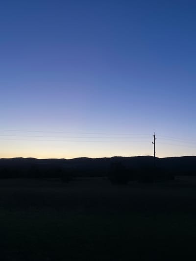 The image depicts a dusky sky transitioning from blue to a faint yellow near the horizon. A power line runs vertically across the scene. Silhouetted mountain ridges are visible along the right side of the frame, giving way to a dark, indistinct area that could be vegetation or terrain. The location is Gorna Kremena, Bulgaria.