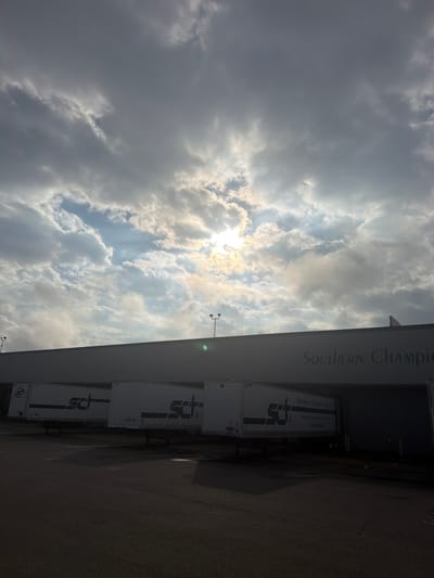 The image captures a low-angle view of a commercial building facade with the name "SOUTHERN CHAMPION" visible in green lettering across the top. Several white semi-trailer trucks are parked in front of the building, some bearing the "SCT" logo. The sky above is filled with dramatic clouds, with the sun partially obscured and shining through, creating a bright effect. The scene appears to be an industrial or logistics area, possibly a distribution center or warehouse. The ground in front of the building is paved, and streetlights are visible on poles. The overall lighting suggests it could be early morning or late afternoon, given the low sun angle and the contrast between the bright sky and the darker foreground.