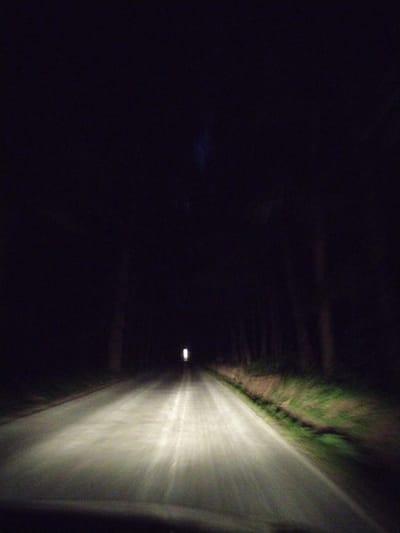 The image depicts a dark, paved road illuminated by headlights. Trees line the sides of the road, creating a sense of being in a wooded area. The sky is not visible, suggesting it is nighttime. The road appears to be in a rural setting, possibly outside of a populated area. There is a single distant light visible on the right side of the road, which could be a streetlight or a distant house. The image is blurry, indicating movement, likely from a vehicle driving on the road. There are no people or other identifiable subjects in the image. The environment is dark and natural, with the road being the only artificial element.