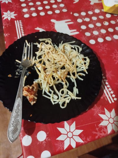 A black plate of spaghetti and a fork rest on a red tablecloth with a white snowflake pattern. The spaghetti is tossed with a light-colored sauce and appears to have some small pieces of meat mixed in. A single piece of meat, covered in red sauce, is on the fork. The setting appears to be indoors, possibly a dining room, given the tablecloth and the presence of a plate of food. The lighting suggests it could be daytime, but it's difficult to be certain without more context. No people are visible in the image. The overall impression is of a simple meal, perhaps after it has been partially consumed. The image is taken from a slightly overhead angle, focusing on the plate of food.