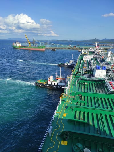 The media file captures a vibrant maritime scene at a port in Kunak, Malaysia, under a bright, clear blue sky with some scattered white clouds, indicative of a sunny day, likely midday or early afternoon.

The dominant subject in the foreground and right side is a large, green-hulled tanker ship, possibly an oil or chemical carrier, with extensive industrial piping, gangways, and structures visible on its deck. This vessel is either docked or in the process of docking alongside a long pier that stretches across the middle ground of the image. A smaller, white and green tugboat with black and yellow markings is positioned near the stern of the large tanker, actively creating a wake in the blue-green water, suggesting it is providing assistance with maneuvering or mooring.

Further along the pier, towards the left of the image, another cargo ship is visible. This vessel has a red hull and a green upper structure, likely a bulk carrier, and appears to be docked. A large yellow gantry crane, part of the port's cargo handling infrastructure, extends over this ship, implying ongoing loading or unloading operations. In the distant background, mountains rise, creating a hazy horizon. The water is relatively calm, reflecting the clear sky and showing ripples from the tugboat's movement.

Several pieces of text are visible on the large green tanker: "NO SMOKING" and "SAFETY FIRST" signs are prominent. Other labels include "KEEP OUT", "PLAY ACCESSIBLE", "ANNEX II MARPOL", "WASTE OIL", and "WR 7P", alongside other technical markings like "BMT" or "BWT". The scene depicts active commercial shipping operations within a harbor environment.