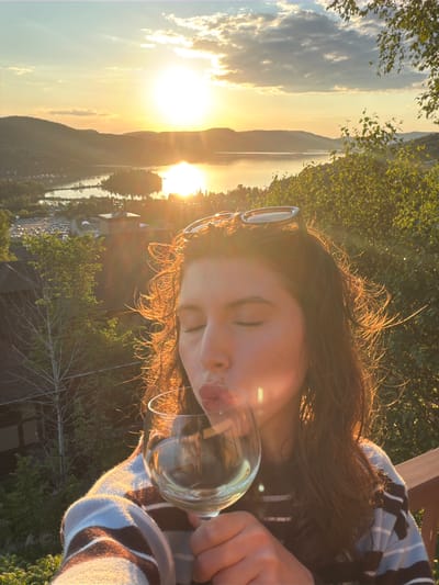 A young woman is captured in a close-up selfie at sunset, holding a glass of white wine. She is wearing a striped sweater and has sunglasses perched on her head. Her eyes are closed, and her lips are pursed as she sips from the glass. The background reveals a picturesque view of Mont-Tremblant, Canada, with rolling hills, a tranquil lake reflecting the golden hues of the setting sun, and a cluster of buildings nestled in the valley. The scene is bathed in the warm, soft light of dusk, creating a serene and idyllic atmosphere.