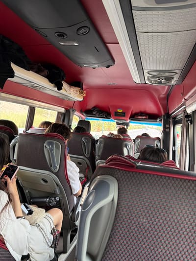 The image depicts the interior of a bus, likely a tour bus, filled with passengers. The bus has red interior walls and ceiling. Overhead compartments run along both sides of the bus. The passenger seats are upholstered in a gray and red patterned fabric. Passengers are seated in the rows, some appearing to be asleep or resting. One passenger is holding a phone. A water bottle and small purse are also visible in the foreground. The bus is moving, as evidenced by the scenery visible through the windows, which includes green vegetation and buildings. The location in Ouarzazate, Morocco cannot be confirmed from the image alone.