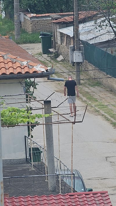 A bald man wearing a black t-shirt and grey shorts walks away from the camera on a paved road in Osetenovo, Bulgaria. He is wearing flip-flops. In the foreground, there is a rusty metal structure with some green foliage growing on it. Behind the man, there are several buildings with corrugated metal roofs and some stone walls. A green trash bin is visible on the left side of the road. Further in the background, there is more vegetation and a utility pole with an electrical box. A dark-colored car is partially visible on the right side of the image. The scene appears to be in a residential or semi-rural area.
