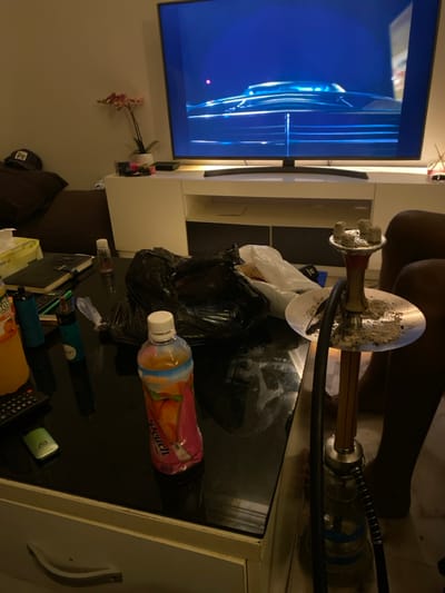 A cluttered living space features a turned-on television with a blue screen. A hookah pipe dominates the central space, resting on a reflective table amidst trash bags and a "peach" flavored beverage bottle. Other items, including a remote, vapes, and what appears to be tissues or napkins, are scattered on the table's surface. The scene has a slightly untidy and indoor feel.