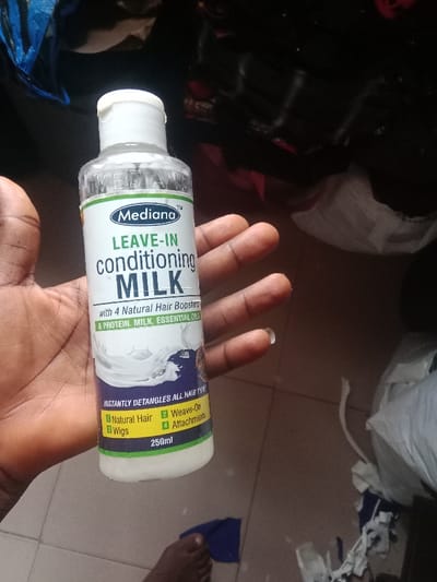 A person holds a clear plastic bottle of Mediana Leave-In Conditioning Milk. The label indicates it contains four natural hair boosters, protein, milk, and essential oils. The bottle claims to instantly detangle all hair types and is suitable for natural hair, weaves, wigs, and attachments. The product volume is 250ml. The person holding the bottle has brown skin. The background is cluttered with clothing and an indistinct floor.