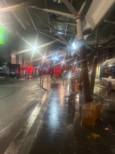 A street scene at night, likely after rain, as the pavement is wet and reflective. The image is taken from under a glass and steel structure. The perspective is from low to the ground, looking down a straight street. There are wooden benches with graffiti on the ends along the right side of the street. Lights shine intensely, casting starburst-like patterns. Red traffic lights are visible in the distance. A car is parked.
