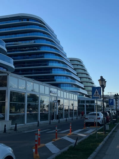 The image depicts a modern, multi-story building with a distinct curved architectural design featuring balconies with blue glass railings. In front of this building, at street level, is a commercial space with large glass windows, possibly a restaurant or shops, as indicated by the text "JUMEIRAH" visible on one of the glass panes. The street in front is paved, with a pedestrian crossing marked by white stripes and orange and white traffic cones. Several cars are parked along the right side of the street, and a street lamp with an ornate design stands next to a pedestrian crossing sign. The sky is clear and blue, suggesting it is daytime, likely late afternoon given the lighting. The scene appears to be a well-maintained urban environment, possibly along a waterfront or promenade, given the sleek, contemporary design of the building and the presence of outdoor seating areas implied by the glass-fronted establishments. The building's design is reminiscent of waves or cascading structures. In the background, construction cranes are visible, indicating ongoing development in the area. The overall impression is one of a modern, possibly resort-style, location in Năvodari, Romania.