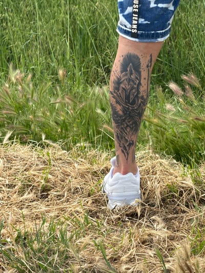 A person's lower leg and foot are positioned between green and dry grass. The person is wearing denim shorts with "USE JEANS" printed on the back, and white sneakers. A detailed tattoo is visible on their lower leg. The location is possibly Kolarovo, Bulgaria.