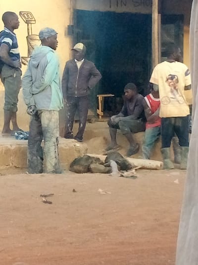 The image depicts an outdoor scene featuring a group of seven men gathered in front of a light yellow building under natural daylight. The ground is a reddish-brown, dusty earth, and the overall setting appears to be a non-urban or commercial area.
On the left side of the frame, three men are standing. The man furthest to the left wears a blue and white striped polo shirt and dark trousers, appearing to be barefoot or in sandals. He is looking towards the right. Adjacent to him, a man is seen from the back, wearing a light blue or grey hooded jacket and dark pants that are heavily soiled with what looks like dirt or cement. He is also looking towards the right. To his right, another man stands with his hands on his hips, wearing a dark jacket over a white shirt and a light-colored baseball cap. His dark trousers also show signs of dust or dirt.
The background features the yellow building, which has a dark, open doorway or entrance in the center. Above this entrance, faint text is visible, reading "TIN SHOP". To the left of the doorway, a simple, hand-drawn illustration of a traditional weighing scale is visible on the building's wall.
In the mid-ground, to the right of the doorway, a man is seated on a low platform or step. He wears a dark long-sleeved shirt and dark pants, both heavily soiled. He is looking downwards or forward. Partially obscured next to him is a younger individual or boy wearing a red vest or shirt. Further to the right, a man stands with his back largely to the camera, wearing a yellow t-shirt that features a graphic design of a person on a bicycle on its back. He is wearing dark pants or shorts and tall, light green rubber boots. On the far right edge of the frame, a small portion of another person is visible, partially obscured by a white cloth or structure in the immediate foreground.
Scattered on the dusty ground in front of the seated and standing figures are several dark rocks and some lighter-colored debris, possibly pieces of paper or fabric. The soiled clothing of many individuals suggests manual labor or activities in a dusty/muddy environment. The men appear to be gathered casually, possibly taking a break or socializing. The lighting indicates it is daytime.