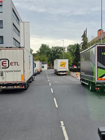The image shows a line of parked trucks on a paved road, possibly in a logistics area or industrial park. The trucks are of various sizes and brands, including "ETL Euro Logistik GmbH" and "Global Logistics" (with a "Geis" logo). One truck is dark green with a striking geometric design and the text "TRAN-STONE LOGISTICS". The road has white lane markings. In the background, there is a modern building with horizontal silver paneling and blue-tinted windows, as well as trees and a cloudy sky. The license plate on the first truck visible reads "F ME 245". A small building with red umbrellas is visible further down the road, suggesting a possible rest area or cafe. The scene appears to be a typical day at a logistics hub, with trucks waiting or parked. The overall impression is one of commercial activity and transport.