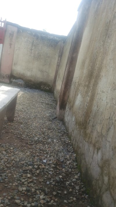The media file shows an outdoor scene in Balewa, Nigeria. The primary subjects are two textured concrete walls forming a corner, with a gravel-covered ground. One wall is lighter in color and appears older, with some dark staining and moss near the base. The other wall is darker and more weathered, with vertical reddish-brown concrete supports or embellishments. The ground is covered in small, unevenly sized gray and brown stones, with patches of darker soil visible. In the foreground left, a portion of a light-colored concrete bench or structure is visible, its edge sharp and clean.
The scene suggests a quiet, possibly neglected outdoor space. There is no activity or event taking place; it appears to be a static view of an architectural setting. Notable details include the texture and condition of the walls, the rough gravel ground, and the presence of the concrete bench. The lighting suggests it is daytime, possibly overcast, as the light is diffused and there are no strong shadows. The location cue "Balewa, Nigeria" provides geographical context. There is no visible text in the image.