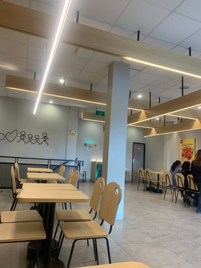 The image depicts the interior of a casual dining establishment located in Calukan RT 1 RW 5 Keboansikep, Indonesia. The space is well-lit by numerous overhead LED strip lights and recessed spot lights integrated into a white grid ceiling, which also features decorative light-toned wooden beams with embedded linear lighting. The flooring consists of light-colored square tiles.
In the foreground and midground on the left, an unoccupied dining area is visible. It features several square tables with light-colored wooden tops and black metal bases, arranged in rows. Each table is accompanied by chairs constructed with light-colored wooden backs, featuring a distinctive circular cutout, and matching seats on black metal frames. This section is partially separated by a low black metal railing. The adjacent wall displays a black stick-figure mural depicting a family (two adults, two children, and a baby) alongside a heart shape.
A prominent light-colored, square structural pillar stands centrally in the midground, extending from floor to ceiling.
In the background on the right, another dining section contains patrons. Four individuals are visible, seated at two separate tables. Their backs are primarily oriented towards the camera. A fifth individual, partially obscured, stands at the far right. The far wall features a large yellow poster with red text and an image of fried chicken, bearing the apparent branding "TIM TIM CHICKEN". A dark brown door is situated on the right wall.
Other objects observed include a red fire extinguisher mounted on a wall to the left of the central pillar, two waste receptacles (one labeled "ORGANIC") in the left background, and a green exit sign with a white arrow positioned high on a wall in the mid-background.