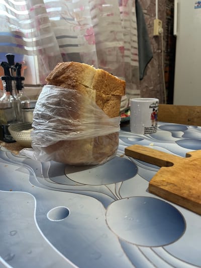 The image captures an indoor scene, likely a kitchen or dining area. A loaf of bread, partially wrapped in clear plastic, is the central subject, positioned on a table with a patterned surface. To the left of the bread, there are several glass bottles and a small bowl containing a white granular substance, possibly salt. A white mug with a printed image and text is visible to the right of the bread. A wooden cutting board lies on the table in the foreground. In the background, a window with sheer curtains and a wall with what appears to be a light switch and a dark cloth hanging are present. The overall lighting suggests a day time setting. There is no indication of the specific location within Altai, Kazakhstan.