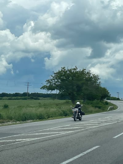 A landscape scene in Buinovo, Bulgaria, shows a winding asphalt road marked with white lines, flanked by green grassy verges and trees. A motorcycle with a rider wearing a white helmet is traveling on the road. The sky is overcast with a mix of white and dark gray clouds, and electrical power lines stretch across the scene.