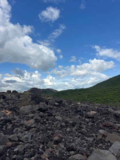 The image shows a rocky, volcanic landscape under a bright blue sky with fluffy white clouds. In the foreground, a dense field of dark, rough-hewn rocks fills the lower portion of the frame. Beyond the rocks, rolling green hills rise, covered in lush vegetation. In the distance, a dark, conical peak, possibly a volcano, is visible against the horizon. The scene appears to be an outdoor natural environment, suggesting a location like the volcanic plains or slopes near Rota, Nicaragua. There are no people or animals present in the image, and no specific actions or interactions are depicted. The overall impression is one of rugged, natural beauty and geological activity.