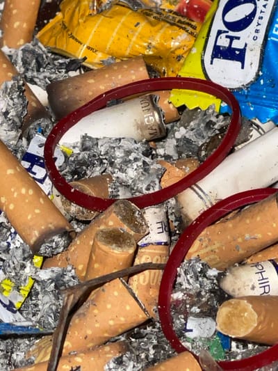This image presents a close-up, overhead view of refuse accumulated within a confined space in Kumbang, Indonesia. The scene is dominated by numerous discarded cigarette butts, primarily featuring tan-orange speckled filters and burnt white paper sections, interspersed with substantial quantities of grey and black ash. Prominently visible objects include one white cigarette butt branded "MAGNUM" on its filter, and another partially visible white filter displaying "SAPPHIR". Two red elastic bands, one larger than the other, are integrated into the debris. Additional items include crumpled packaging fragments: a yellow foil wrapper with partial text indicating "Indonesia," and a blue and white foil wrapper displaying "FO" and "CRYSTAL." A thin, dark, stick-like object is also present. No individuals are visible in the image.
