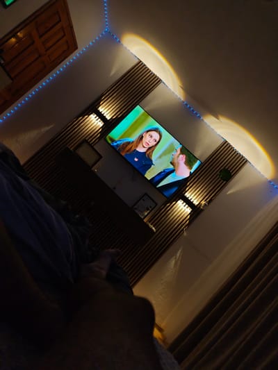 The image depicts an indoor scene in a bedroom, likely in Grohotno, Bulgaria. A large flat-screen television is mounted on a wooden slatted wall, displaying a scene with two individuals, a woman with blonde hair and a man with dark hair. The room is dimly lit, with blue LED strip lights outlining the ceiling and a warm gradient light casting an arc on the wall above the television. To the left of the television, a wooden door with paneling is visible. The foreground is dominated by the dark silhouette of a person lying down, covered by a blue blanket or sheet. The scene suggests a relaxed atmosphere, with the person in the foreground watching television.