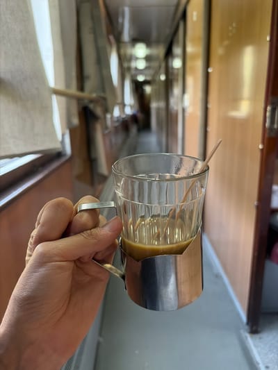 A hand holds a clear glass mug with a thin, light-brown liquid inside. A thin wooden stirrer protrudes from the mug's opening. The mug is encased in a polished silver holder with a handle. The background is blurred and shows a long, narrow interior space with wooden paneling and overhead lighting. The perspective suggests the viewer is looking down a corridor or aisle. The lighting appears to be artificial, possibly from indoor lights. The overall impression is of an interior setting, possibly a train car or a long room, with a beverage being prepared or served. There is no visible text.