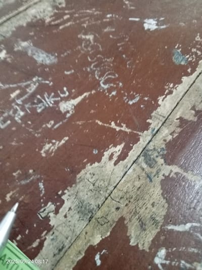 A close-up, oblique view depicts a heavily worn and defaced wooden surface, likely a desk or tabletop, located in Quebrada del Medio, Venezuela. The surface, predominantly reddish-brown, exhibits significant deterioration of its finish, with extensive areas of chipping, flaking, and abrasion revealing lighter, possibly natural wood beneath. Numerous light-colored scratches, carved lines, and partial alphanumeric characters, including what appears to be "C", "K", "U", and "S", are etched across the surface. A prominent dark linear seam or crack runs diagonally through the lower right quadrant. In the lower-left corner, the silver-colored tip of a mechanical pencil is visible, resting on or adjacent to the surface, partially obscuring a sliver of green material. A digital timestamp "2025/09/24 08:17" is overlaid in the bottom-left corner. No individuals are present in the scene.