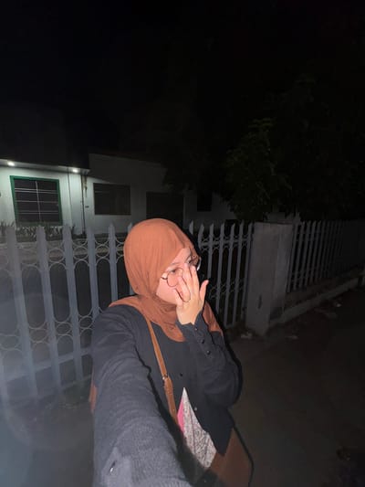 In the city of Tanggul, Indonesia, at night, a person wearing a brown hijab and glasses is taking a selfie. They are also wearing a black cardigan over a floral-patterned shirt and a brown bag. The background shows a white building with green-framed windows, a white fence, and trees. The scene is dimly lit, with the primary light source appearing to be from the building.