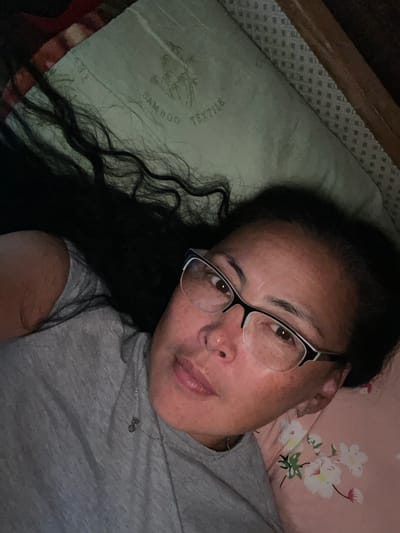 A woman with long, dark, wavy hair and glasses is lying on a bed, looking at the camera. She is wearing a grey t-shirt with a small silver pendant. The bed has a pillow with a pattern of bamboo and the word "TEXTILE" printed on it, and a pink pillow with a floral print. The scene appears to be indoors, possibly in a bedroom. There is no discernible location in the city of Bulgunnyakhtakh, Russia.