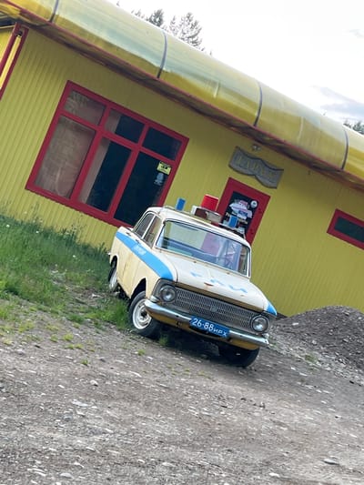 In Bolshaya Rechka, Russia, a vintage, light-colored police car with a blue stripe and a blue and red light bar on its roof is parked on a gravelly incline in front of a bright yellow building. The building has a large red-trimmed window on the left and a red-trimmed door on the right, with a sign above it that reads "ГАРАЖ" (GARAGE). The car's license plate is visible, reading "26-88 ирх". The ground is covered in gravel and sparse grass. In the background, trees are visible against a cloudy sky. The scene appears to be outdoors.