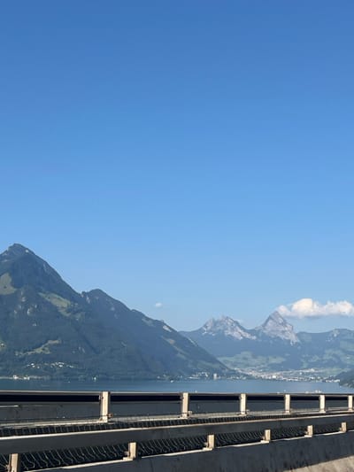 The image displays a panoramic outdoor scene from an elevated vantage point, likely a bridge or highway, offering a view across a large lake towards a distant mountain range under a clear sky. The scene is located near Beckenried, Switzerland, looking across Lake Lucerne (Vierwaldstättersee) towards the north-northeast.
In the foreground, occupying the bottom approximately 20% of the frame, is a gray concrete barrier or railing structure. It features a solid base topped by two parallel horizontal metal rails supported by several light-colored vertical posts evenly spaced across the width of the image. A dark, possibly mesh-like, material is visible between the rails.
Beyond the barrier, the midground is dominated by the calm, dark blue waters of a large lake, extending horizontally across the frame. Small, indistinct white specks are faintly visible on the water's surface. On the far shore, nestled at the base of the distant mountains, a cluster of buildings and structures indicates a small town or settlement.
The background is defined by a majestic mountain range. The mountains closest to the left are covered densely with green foliage and appear to rise steeply from the lake's edge. Further to the right and in the distance, more rugged, rocky peaks emerge, several displaying patches of lighter-colored rock or snow near their summits, including two prominent, jagged peaks. The atmospheric perspective renders the most distant peaks slightly hazy.
The upper two-thirds of the image are filled with a clear, vivid blue sky. A few small, wispy white clouds are clustered low on the horizon towards the right side of the frame. No individuals, animals, or discernible actions are present within the scene.