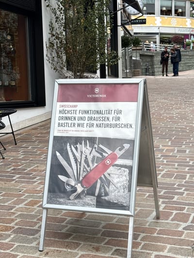 An outdoor advertising stand displays a poster for Victorinox Swiss Army knives. The poster features a large red Swiss Army knife surrounded by various tools and blades against a black and white background. The text on the poster, in German, reads "SWISSCHAMP HÖCHSTE FUNKTIONALITÄT FÜR DRINNEN UND DRAUSSEN, FÜR BASTLER WIE FÜR NATURBURSCHEN." Below this, it says "FROM THE MAKERS OF THE ORIGINAL SWISS ARMY KNIFE™ ESTABLISHED 1884". The advertising stand is positioned on a cobblestone street. In the background, there are several people walking and interacting. Buildings with shops are visible, including one with signage that reads "HAWICO" and another with "PIZZERIA". The scene appears to be in a pedestrian area of a European city, likely in Italy given the signage. The overall setting is a commercial street with an urban environment.