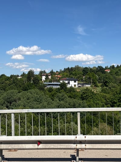 The image depicts a bright, sunny day with a clear blue sky dotted with white, fluffy clouds. In the foreground, a metal guardrail with vertical bars separates the viewer from a lush, green hillside densely covered with trees. Several houses are visible nestled among the trees on the hillside. One prominent house is white with a black roof, featuring large windows. Other houses appear to have red or brown roofs. A solar panel array is visible on a flat-roofed structure near the white house. The scene suggests a residential area situated in a natural, hilly environment. This view is consistent with the location in Malo Buchino, Bulgaria, known for its scenic landscape and scattered residential properties within green surroundings.