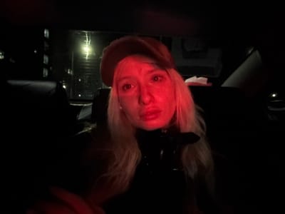 An upside-down, dark, and red-toned image shows a blonde person looking at the camera. They are wearing a dark jacket or top, and a beige hat. In the background, there appears to be a night cityscape with buildings and lights. The lighting is dim, with a strong red hue dominating the scene. The overall atmosphere is mysterious.
