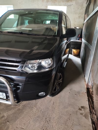 A black van is parked in a garage. The van is facing the viewer, with the front of the vehicle prominently displayed. The garage appears to be made of concrete block and has a corrugated metal wall on the right side. There are several tires stacked in the corner of the garage, and a yellow bucket is also visible. The floor of the garage is concrete and has tire tracks on it. The overall impression is one of a utilitarian space. The image was taken in Barysaw, Belarus.